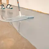 Osoba w butach rozlewa szarą żywicę na betonową podłogę. Widać nierówności, które mogą być wadami podłogi z żywicy.