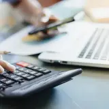 Dłoń z ołówkiem liczy na kalkulatorze, obok laptop i kubek. To pomoc w zrozumieniu, jak obliczyć zużycie prądu.