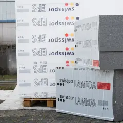 Stosy płyt izolacyjnych z napisem "swisspor LAMBDA" i "EPS" na paletach, gotowe do budowy.