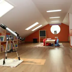 Na bardzo niskim i małym poddaszu stoi teleskop, obok regały z książkami i kącik do zabawy z tablicą.