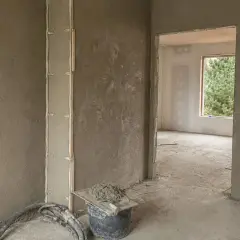 Ściany w trakcie prac wykończeniowych, na których widać świeże tynki gipsowo wapienne. W wiadrze zaprawa i kielnia.