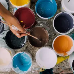 Ręka miesza farby w wiaderkach. Widać różne kolory: żółty, czerwony, niebieski, brązowy, pomarańczowy, czarny, biały. Wady farby ceramicznej mogą objawiać się nierównym kryciem.
