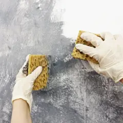 Dwie ręce w białych rękawiczkach wykonują malowanie gąbką na szarej ścianie, tworząc artystyczną teksturę.