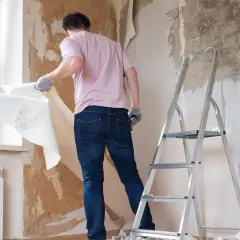 Mężczyzna w rękawiczkach zrywa starą tapetę ze ściany, pokazując, jak usunąć tapetę ze ściany. Obok stoi drabina.
