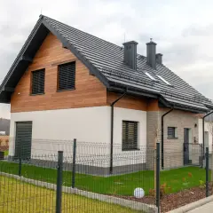 Nowoczesny dom z drewnianą elewacją i czarnym, dwuspadowym dachem. Ogrodzony teren z zadbanym trawnikiem i ozdobnymi kulami.