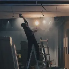Budowa wnętrza, pracownik na drabinie montuje sufit, w tle materiały budowlane i lampy.