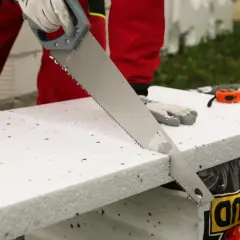 Ręce w rękawiczkach tną styropian zwykłą piłą ręczną. To prosty sposób, czym ciąć styropian.