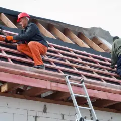 Dwaj budowlańcy pracują na dachu, montując wiatroizolację. Jeden z nich, w pomarańczowych spodniach i czerwonym kasku, wbija gwoździe.