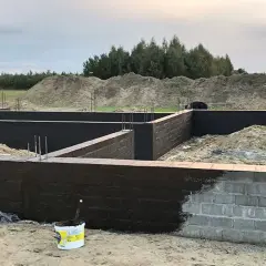 Fundament domu w budowie, ściany fundamentowe pokryte ciemną masą izolacyjną. Trwa izolacja powłokowa.