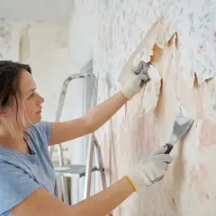 Kobieta w rękawiczkach zeskrobuje starą tapetę, przygotowując ścianę do położenia nowej.