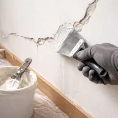 Ręka w rękawicy nakłada masę szpachlową na głębokie pęknięcia w ścianie. Obok wiadro z masą i narzędzia do naprawy.