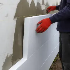 Pracownik w czerwonych rękawicach przykłada styropian do ściany. Dowiedz się, ile kosztuje ocieplenie domu styropianem.
