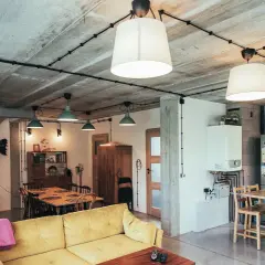 Nowoczesne wnętrze z betonowym sufitem, gdzie widoczna jest **instalacja elektryczna natynkowa** prowadząca do lamp. Widać kanapę, stół i kuchnię.