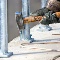 Dłoń w rękawicy uderza młotkiem w metalowy pręt przykręcony do betonu.