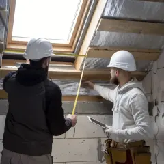Dwóch budowlańców w kaskach mierzy taśmą okno dachowe. Sprawdzają, do jakiej temperatury można kleić styropian, by zapewnić dobrą izolację.