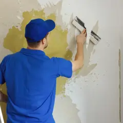 Mężczyzna w niebieskiej koszulce i czapce usuwa starą tapetę za pomocą szpachelki. Prace remontowe, usuwanie farby lateksowej.