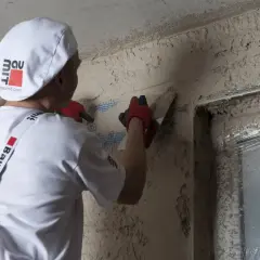 Pracownik w białej czapce i ogrodniczkach wykonuje zacieranie tynku na ścianie, używając kielni.