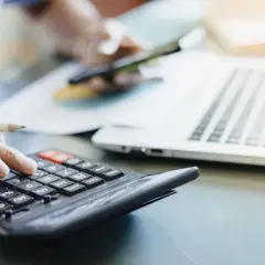 Dłoń z ołówkiem liczy na kalkulatorze, obok laptop i kubek. To pomoc w zrozumieniu, jak obliczyć zużycie prądu.