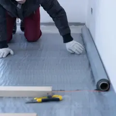 Układanie folii na styropian. Pracownik w rękawiczkach rozkłada szarą folię izolacyjną na podłodze, przygotowując ją pod dalsze prace.