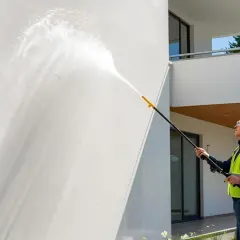 Mężczyzna w kamizelce odblaskowej myje elewację domowym sposobem, używając myjki ciśnieniowej.