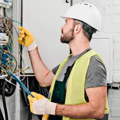Elektryk w kasku i kamizelce odblaskowej pracuje przy skrzynce z przewodami. Zastanawia się, ile za punkt elektryczny zapłaci klient.