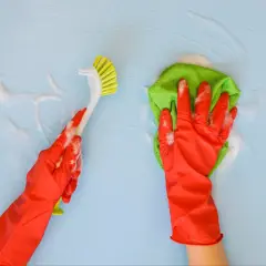 Ręce w czerwonych rękawiczkach myją niebieską ścianę. Jedna ręka trzyma zieloną ściereczkę, druga szczotkę. Dowiedz się, czym myć ściany.
