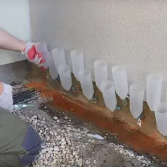 Pracownik w rękawiczkach montuje system do osuszania murów, umieszczając plastikowe rurki wzdłuż ściany.