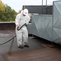 Pracownik w kombinezonie ochronnym wykonuje renowację papy termozgrzewalnej na dachu, natryskując preparat.
