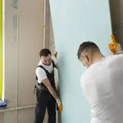 Dwóch budowlańców montuje płytę gipsowo-kartonową. Prace przy zakończeniu płyty gk idą sprawnie.