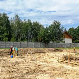 Pracownicy wykonują przygotowanie terenu pod budowę. Widać wykopane fundamenty, geodetę z niwelatorem i koparkę.