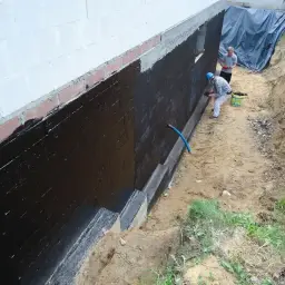 Pracownicy nakładają masę bitumiczną na fundamenty domu, zabezpieczając je przed wilgocią.