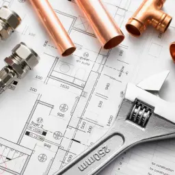 Projekt instalacji wodnej w domu: miedziane rury, złączki, węże i klucz nastawny leżą na planie architektonicznym.