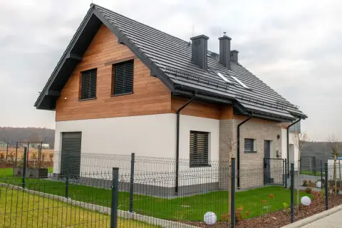 Nowoczesny dom z drewnianą elewacją i czarnym, dwuspadowym dachem. Ogrodzony teren z zadbanym trawnikiem i ozdobnymi kulami.