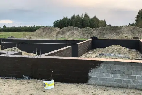 Fundament domu w budowie, ściany fundamentowe pokryte ciemną masą izolacyjną. Trwa izolacja powłokowa.