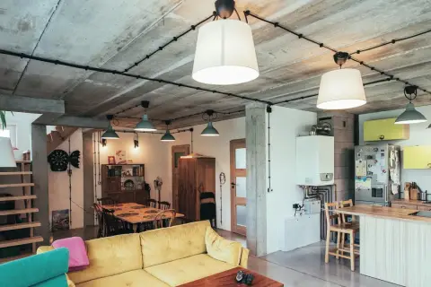 Nowoczesne wnętrze z betonowym sufitem, gdzie widoczna jest **instalacja elektryczna natynkowa** prowadząca do lamp. Widać kanapę, stół i kuchnię.