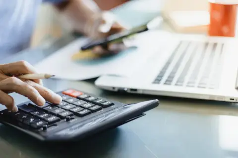 Dłoń z ołówkiem liczy na kalkulatorze, obok laptop i kubek. To pomoc w zrozumieniu, jak obliczyć zużycie prądu.