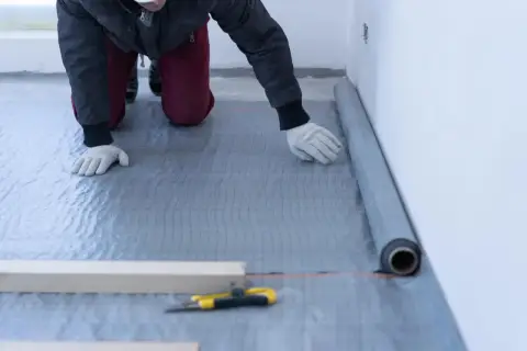 Układanie folii na styropian. Pracownik w rękawiczkach rozkłada szarą folię izolacyjną na podłodze, przygotowując ją pod dalsze prace.