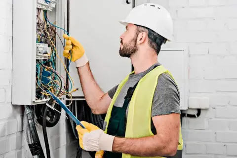 Elektryk w kasku i kamizelce odblaskowej pracuje przy skrzynce z przewodami. Zastanawia się, ile za punkt elektryczny zapłaci klient.