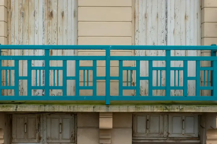 Turkusowe mocowanie balustrady do ściany z ociepleniem, z widocznymi drewnianymi drzwiczkami poniżej.