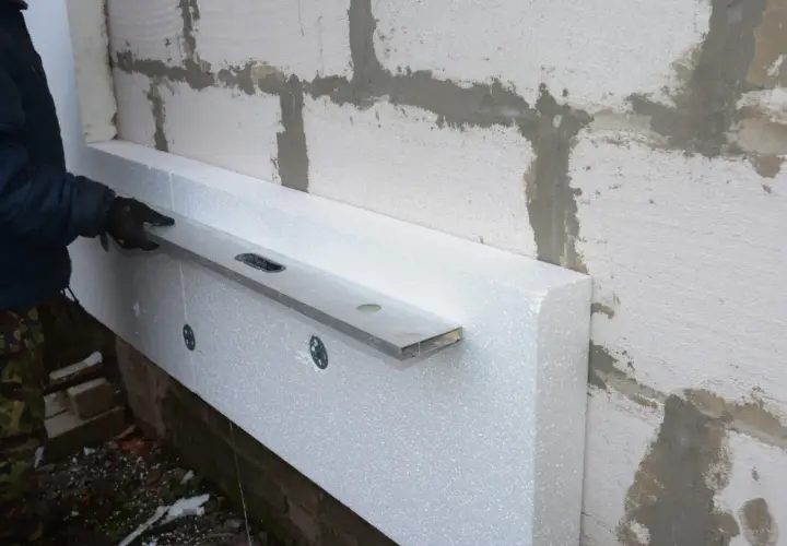 Osoba w rękawiczkach sprawdza poziomicą, jak układać styropian na podłodze, przy ścianie z bloczków.