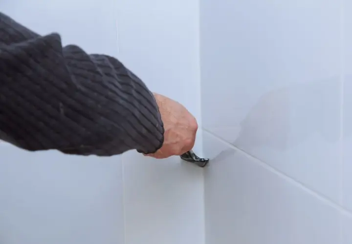 Ręka w swetrze usuwa stary silikon z białych płytek za pomocą narzędzia. Domowy sposób na usuwanie silikonu octem może pomóc w tej pracy.