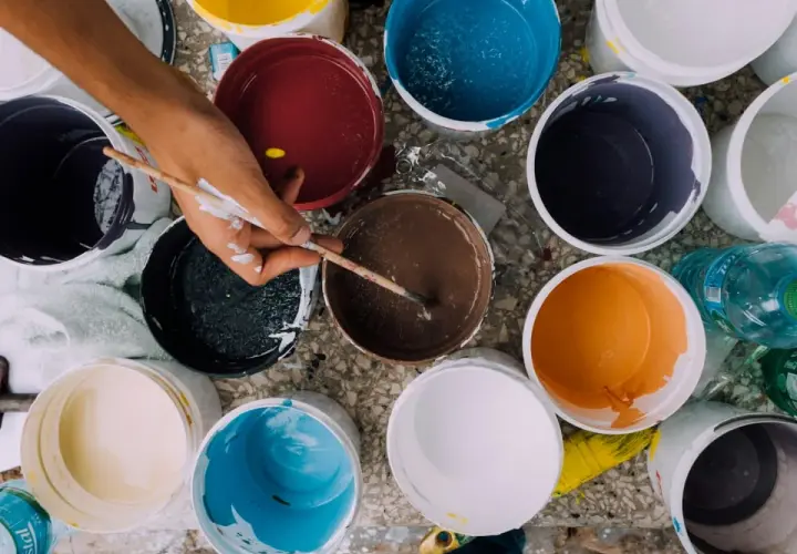 Ręka miesza farby w wiaderkach. Widać różne kolory: żółty, czerwony, niebieski, brązowy, pomarańczowy, czarny, biały. Wady farby ceramicznej mogą objawiać się nierównym kryciem.