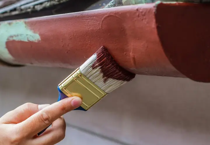 Ręka maluje pędzlem rynny, odnawiając stare malowanie aluminium.