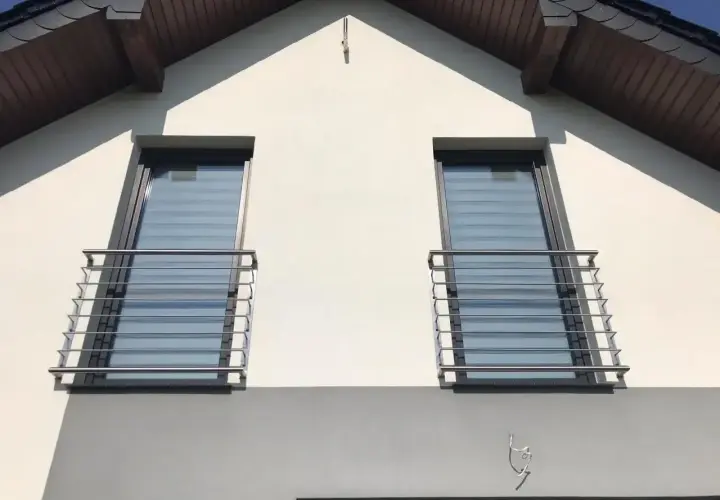 Nowoczesny montaż balkonu francuskiego na białej elewacji domu. Dwa okna z balustradami ze stali nierdzewnej.
