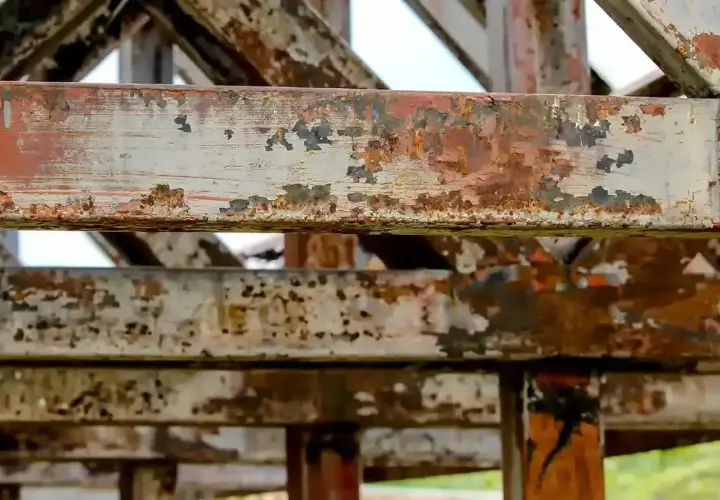 Metalowe belki konstrukcji pokryte łuszczącą się farbą i rdzą. Widać ślady postępującej korozji, która niszczy materiał.