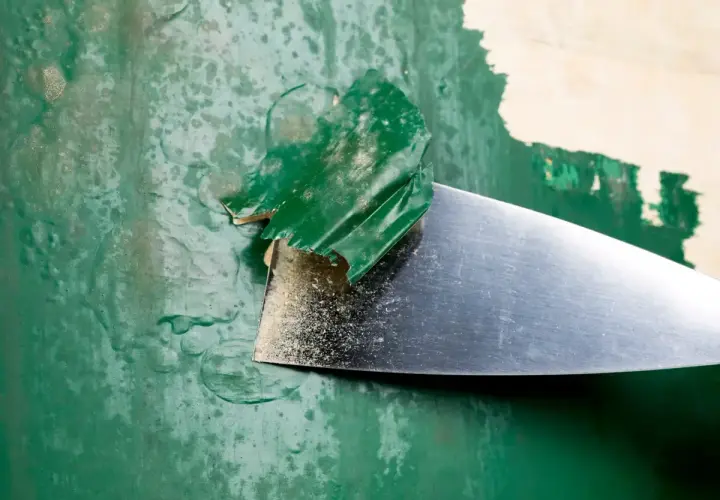 Metalowa szpachelka zdziera starą, zieloną farbę, odsłaniając podkład.