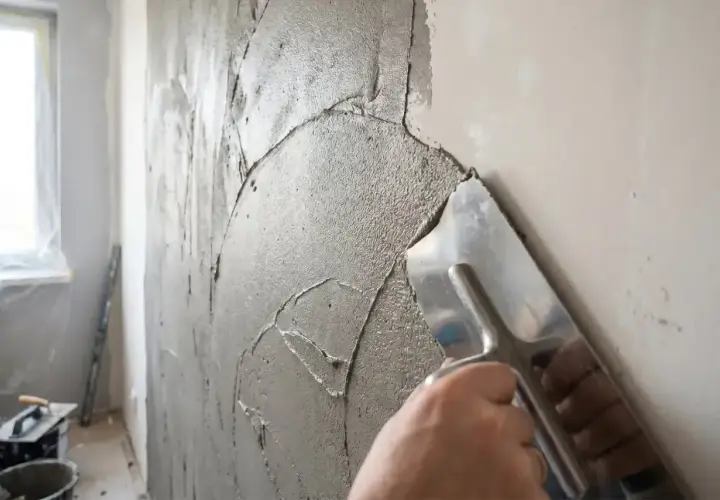 Ręka z kielnią nakłada grubą warstwę masy naprawczej na ścianę, tworząc nierówną fakturę.