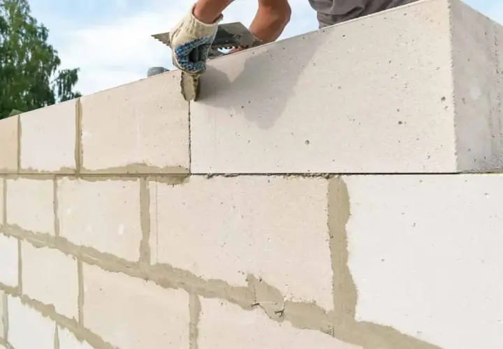 Budowa ściany z bloczków betonowych. Ręka w rękawicy nakłada zaprawę, dbając o zachowanie minimalnej grubości ściany nośnej.