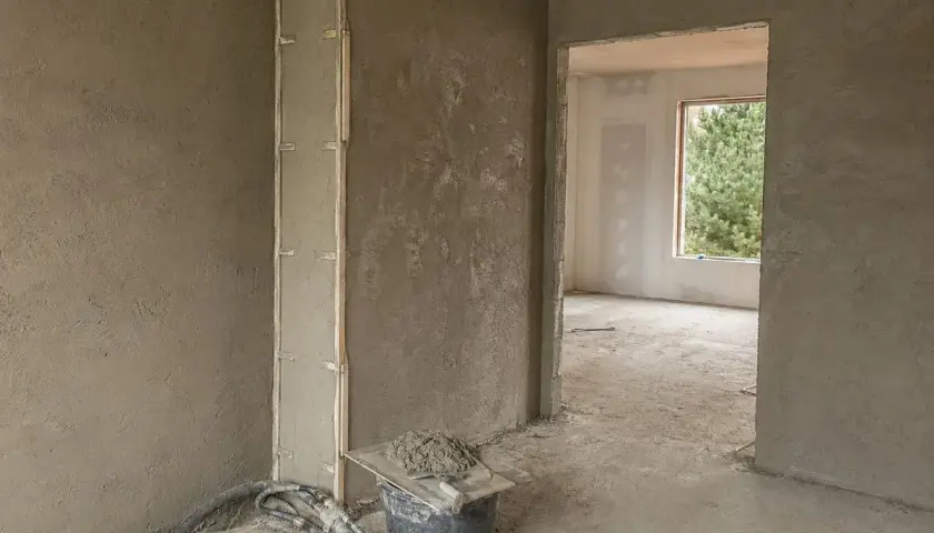 Ściany w trakcie prac wykończeniowych, na których widać świeże tynki gipsowo wapienne. W wiadrze zaprawa i kielnia.