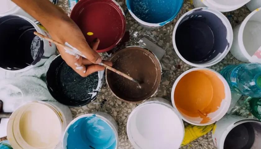 Ręka miesza farby w wiaderkach. Widać różne kolory: żółty, czerwony, niebieski, brązowy, pomarańczowy, czarny, biały. Wady farby ceramicznej mogą objawiać się nierównym kryciem.